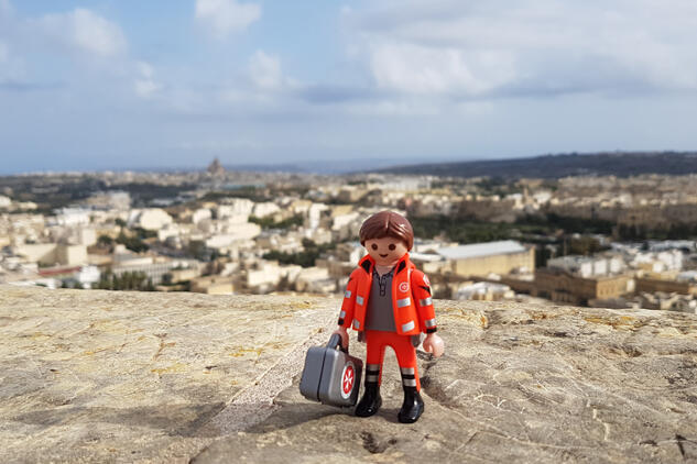 Playmobil-Figur der Johanniter steht in Malta. Im Hintergrund sind das Meer und die tollen Wohnhäuser der Inselbewohner zu sehen.
