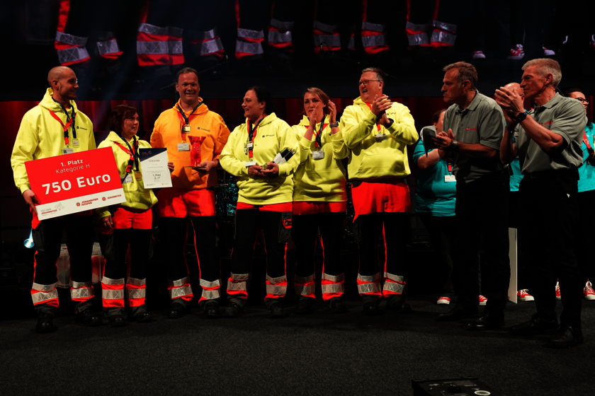 Siegerehrung der Rettungshundestaffel beim Tag der Johanniter 2023 Acht Menschen stehen im Halbkreis auf einer Bühne. Einige klatschen und lachen. Ein Mann hält ein Schild mit der Aufschrift "1. Platz Kategorie H, 750 Euro" in der Hand. Die drei Frauen und drei Männer auf der vom Betrachter aus linken Seite tragen gelbe Hoodues und rot-schwarze Einsatzhosen; die beiden Männer rechts graue Poloshirts.