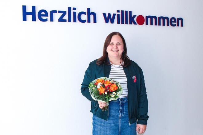 Eine Frau mit Blumenstrauß vor einer weißen Wand mit der Aufschrift "Herzlich willkommen"