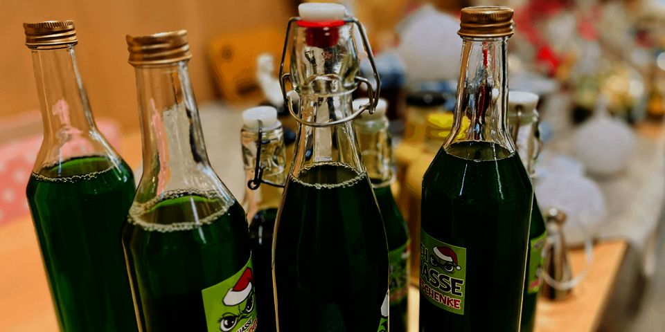 Mehrere Flaschen mit leuchtend grünem Likör, versehen mit satirischen „Grinch“-Weihnachtsaufklebern, stehen dekorativ auf einem Tisch.