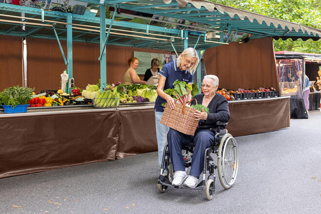Eine Frau reicht einer älteren Frau, die in einem Rollstuhl sitzt, einen Korb mit Gemüse. Sie befinden sich auf einem Wochenmarkt.