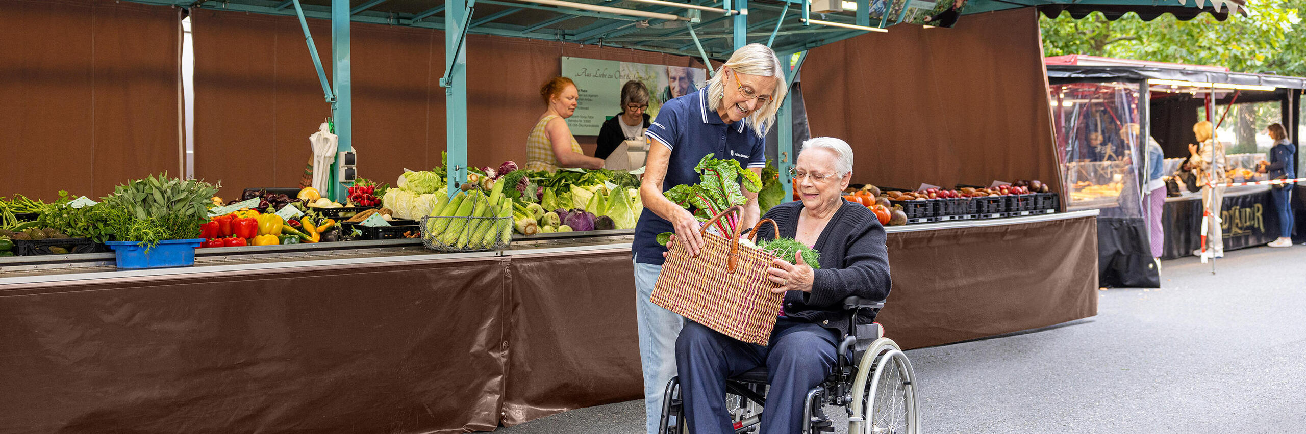 Eine Frau reicht einer älteren Frau, die in einem Rollstuhl sitzt, einen Korb mit Gemüse. Sie befinden sich auf einem Wochenmarkt.