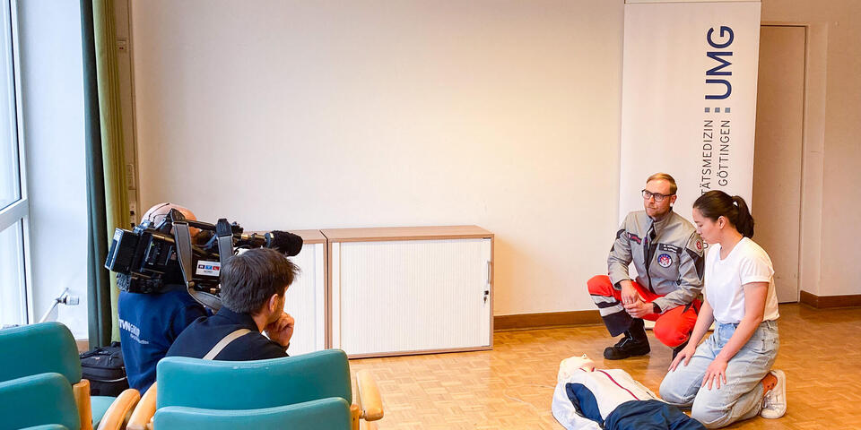 Ein Johanniter und eine junge Frau knien vor einer Reanimationspuppe ein Kamerateam filmt dies.