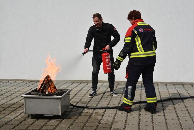 Mann löscht Feuer mit Übungsfeuerlöscher, begleitet von Johanniter-Ausbilderin.