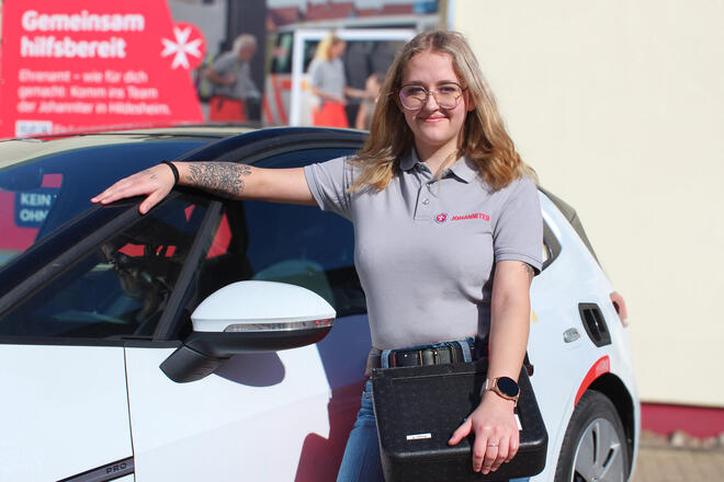 Eine junge Frau mit Johanniter-Poloshirt und einer Menübox vor einem Johanniter-Fahrzeug