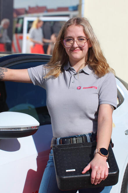 Eine junge Frau mit Johanniter-Poloshirt und einer Menübox vor einem Johanniter-Fahrzeug