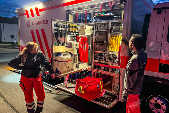 Ein Mann und eine Frau in Einsatzkleidung vor einem großen Gerätewagen. Die Seitentür des Fahrzeugs ist offen und man sieht viele Gerätschaften