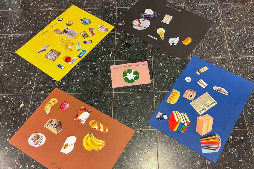 Verschiedenfarbige Pappen liegen auf dem Boden, darauf liegen Bilder von Müllsorten, die Pappen zugeordnet wurden.