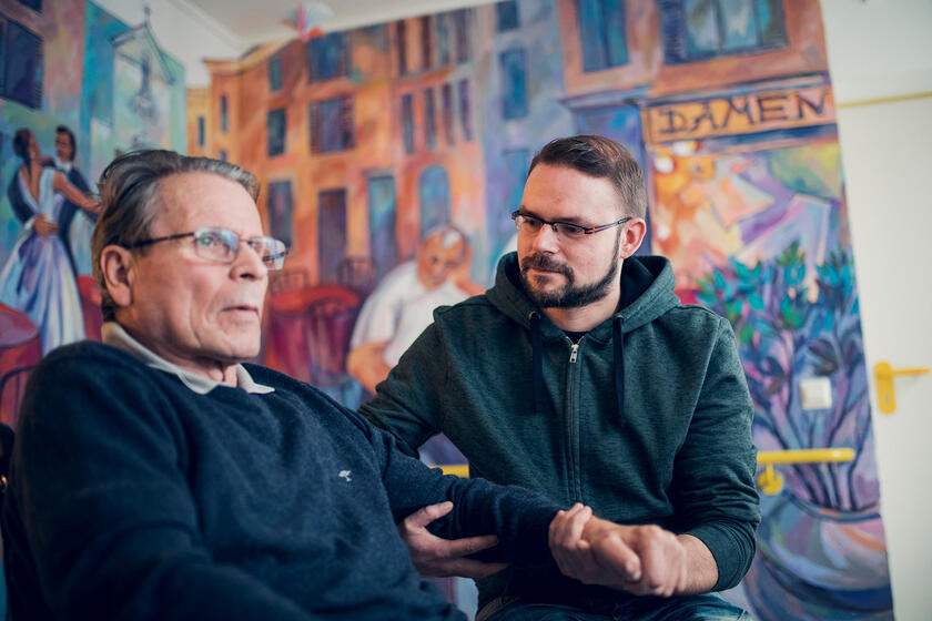 Ein jüngerer Mann im blauen Pullover hilft einem Bewohner des Johanniter-Haus Empelde