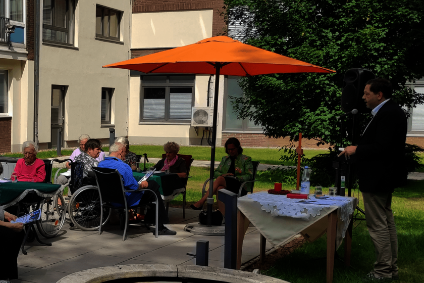 Menschen sitzen im Freien an Tischen. Ein Mann steht hinter einem Tisch, auf dem ein weißes Tischtuch liegt.