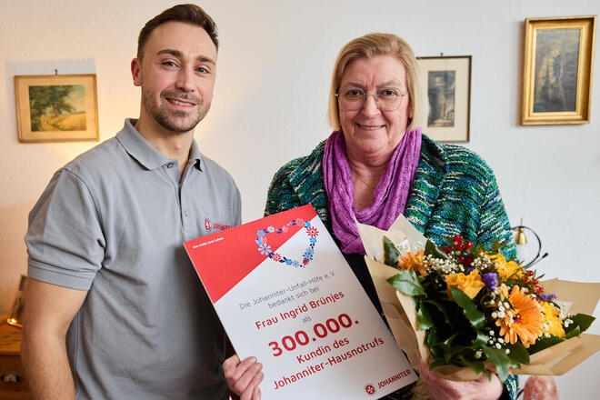 Ein Mitarbeitender der Johanniter überreicht der 300.000 Hausnotruf-Kundin der Johanniter in ihrer Wohnung einen Blumenstrauß und eine Dankesurkunde der Johanniter.