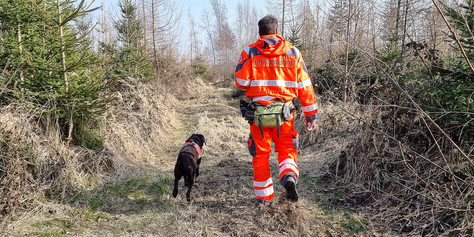 Ein Johanniter der Rettungshundestaffel läuft mit seinem braunen Hund über einen Weg zwischen Bäumen