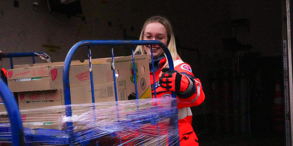 Eine Frau in Johanniter-Kleidung schiebt einen gefüllten Rollbehälter.