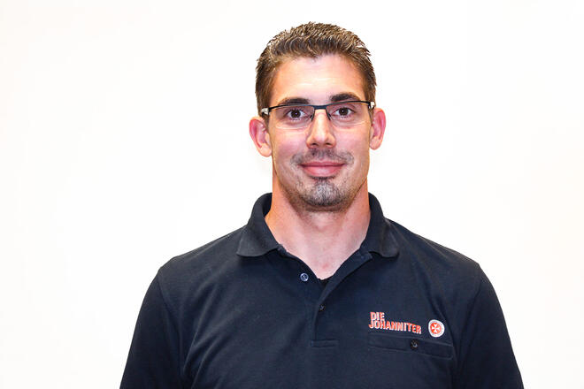 Ein Mann mit braunen Haaren und einem Kinnbart und Brille im dunkelblauem Poloshirt