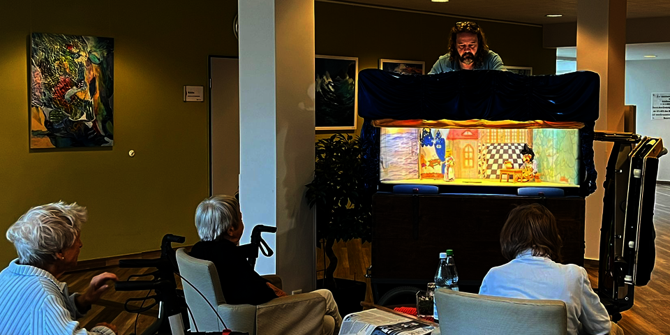 Ein Mann steht gebückt hinter einem größeren Kasten, in dem eine erleuchtete kleine Bühne ist. Drei zuschauende Seniorinnen sind von hinten zu sehen.