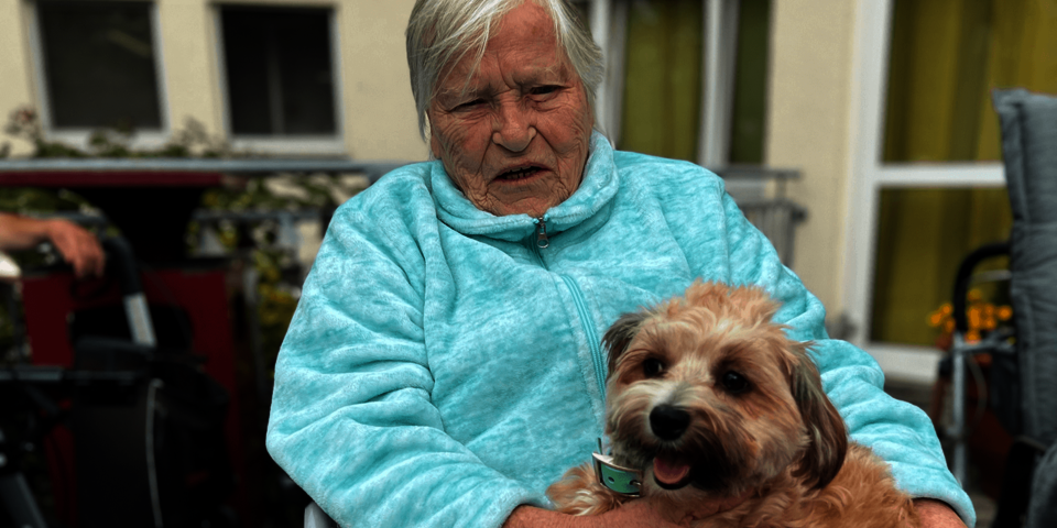 Eine Frau mit weißen Haaren trägt eine Jacke in Türkis. Sie sitzt in einem Rollstuhl und hält einen kleinen Hund auf dem Schoß.