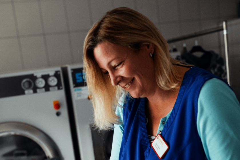 eine lächelnde blonde Frau in türkisem T-Shirt und blauem Kasack vor einer Reihe Waschmaschinen