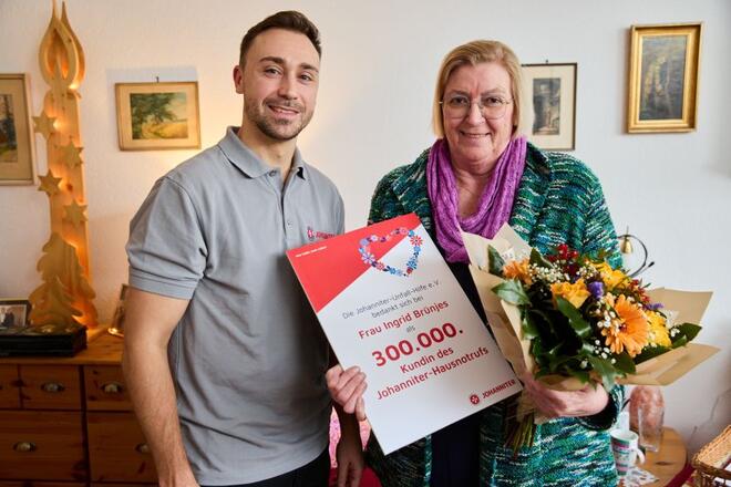 Eine ältere Person und ein jüngerer Mitarbeitender der Johanniter stehen lächelnd nebeneinander. Die ältere Person hält einen Blumenstrauß und eine Urkunde in den Händen. Auf der Urkunde ist die Zahl „300.000“ zu sehen. Die Stimmung wirkt feierlich und freundlich; das Foto vermittelt Wertschätzung, Sicherheit und persönlichen Service.