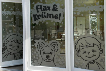 3 Fensterscheiben beklebt mit Comicfiguren, links ein Mädchen, in der Mitte ein Hund, rechts ein Junge und alle drei stehen für den Kitanamen Flax & Krümel
