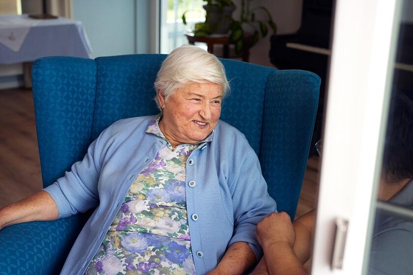 Eine ältere Frau sitzt in einem blauen Sessel am Fenster