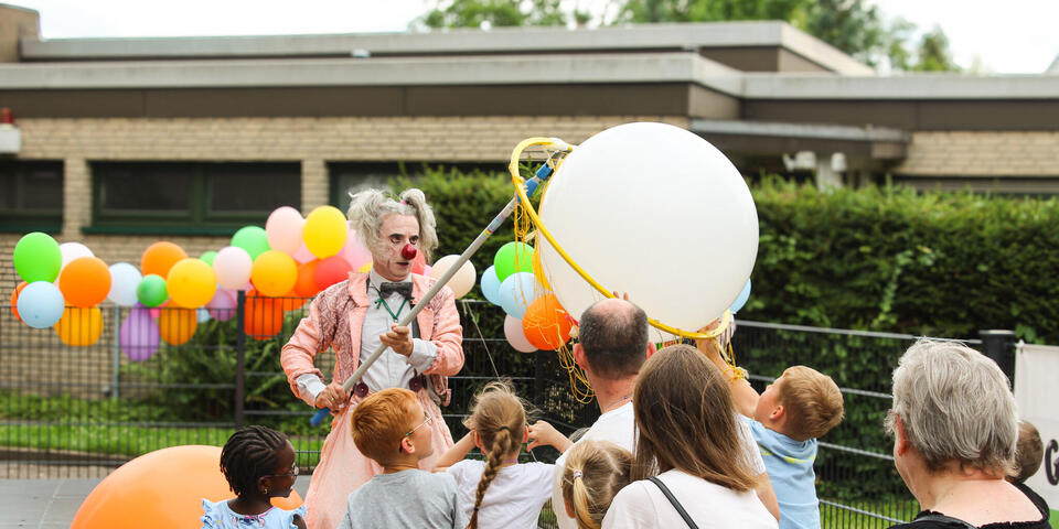 Ein Clown mit einem riesigen weißen Luftballon. Kinder und Erwachsene schauen ihm zu.