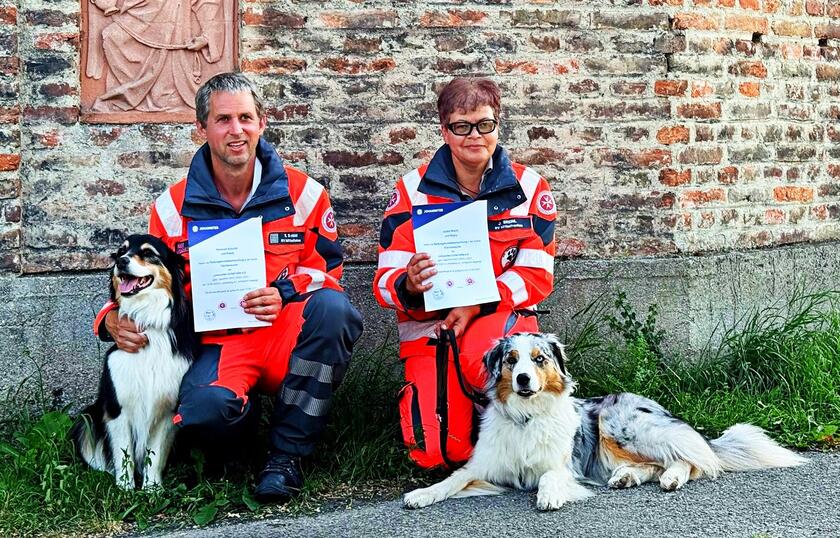 Zwei Personen in Einsatzkleidung der Johanniter sitzen vor einer Backsteinmauer, halten Urkunden und sind jeweils mit einem Suchhund zu sehen.