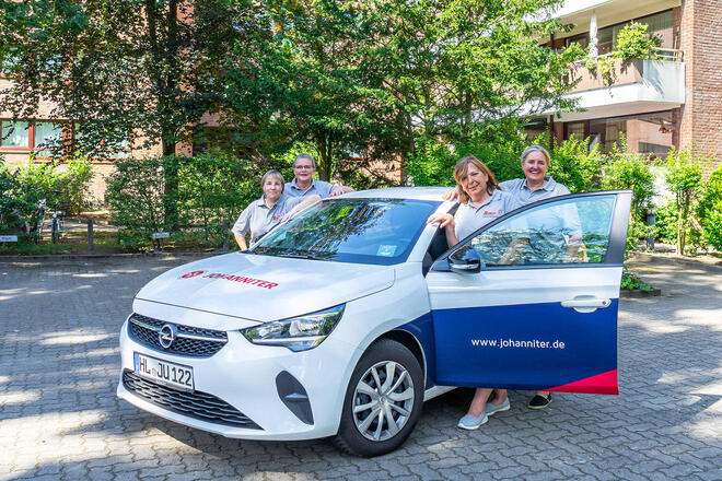 Die Pflegedienstmitarbeiterinnen stehen um ein Auto des Ambulanten Pflegedienstes und lächeln in die Kamera.