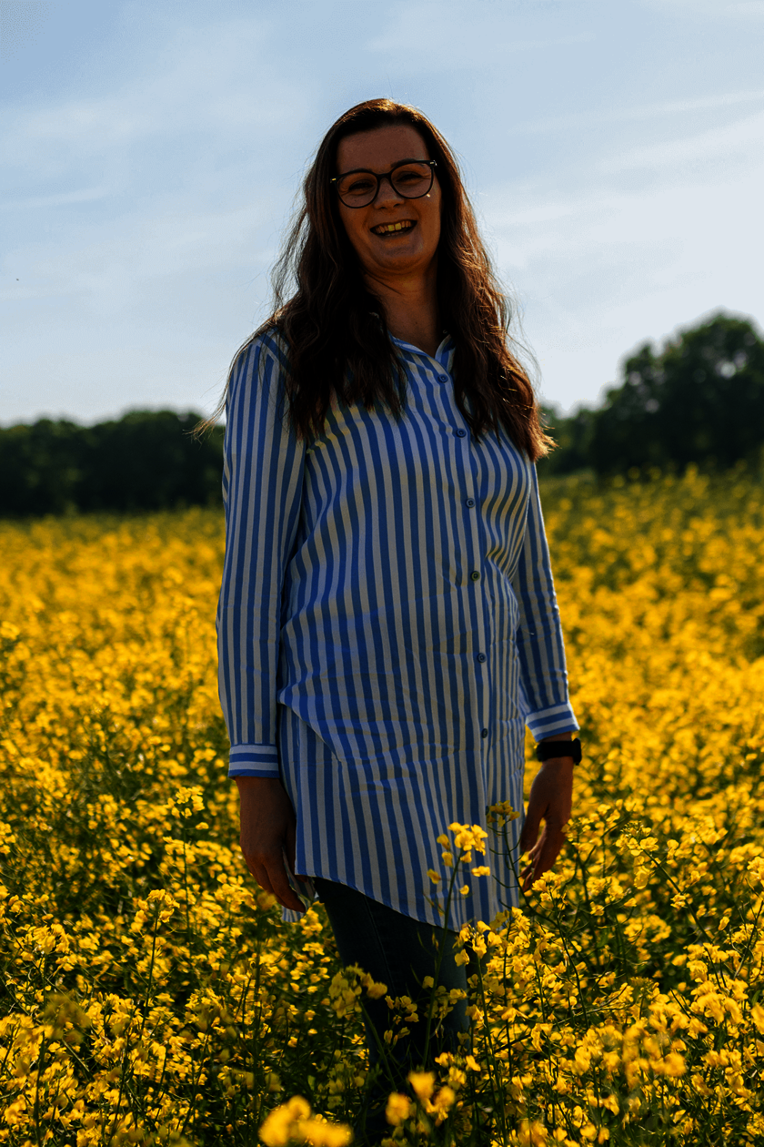 Frau steht in einem Rapsfeld und lächelt, weite gelbe Blumen im Hintergrund.  