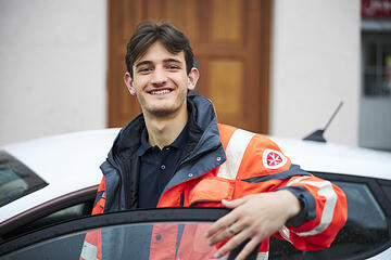 Ein junger Mann in einer Einsatzjacke steht lächelnd neben einem Auto und hält die geöffnete Tür.