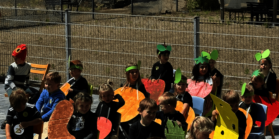 18 Kinder sitzen im Freien auf Stühlen. Sie halten große Pappfiguren in den Händen und tragen zum Teil auf dem Kopf gebastelte grüne Blätter aus Papier.