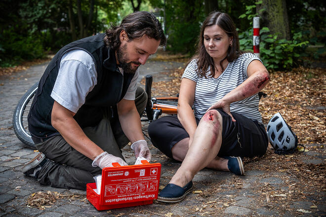 Ein Autofahrer versorgt eine Radfahrerin nach einem Sturz. Sie hat Verletzungen an Armen und Beinen. Der Autofahrer nutzt einen Verbandkasten der Johanniter-Unfall-Hilfe e.V