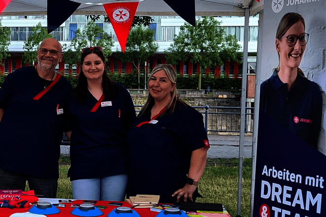Zwei Frauen und ein Mann in dunkelblauen Polos stehen lächelnd hinter einem Stand unter einem weißen Pavillon. Rechts und links stehen Rollups, die für die Arbeit bei den Johanniter werben. Auf dem Tisch vor den Personen liegen zur Deko rote Servietten und verschiedene Gegenstände als Give aways.