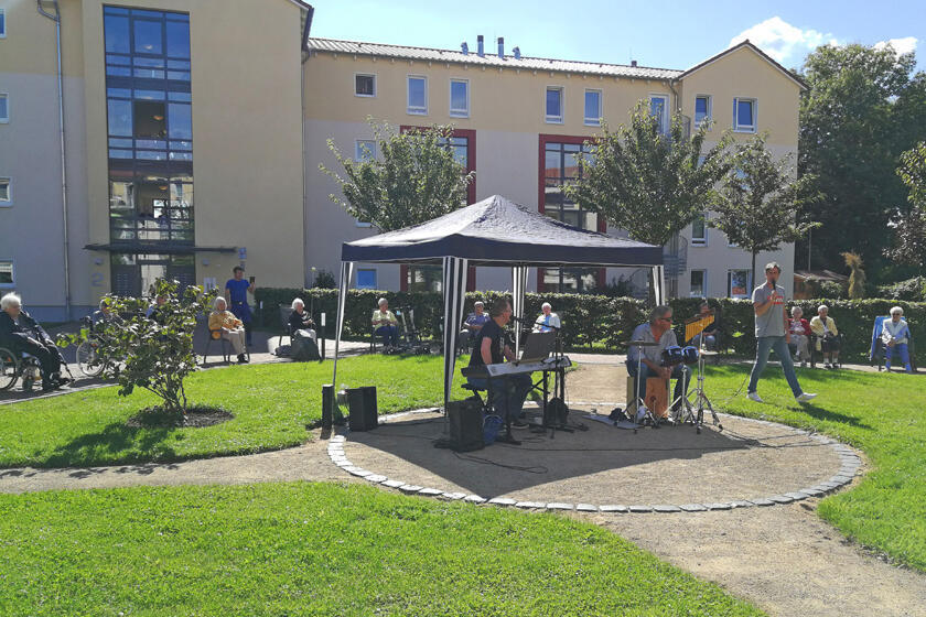 Musiker mit Kiboard und Mikrofone spielen zahlreiche Lieder auf der Wiese. Mit großem Abstand sitzen die Zuhöhrenden im Kreis um die Band herum. 