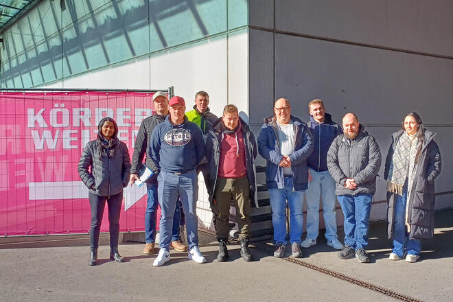 Zwei Frauen und und sieben Männer vor Einer Veranstaltungshalle. Links von ihnen hängt ein großes pinkes Bauzaunplakat auf dem steht "Körperwelten"
