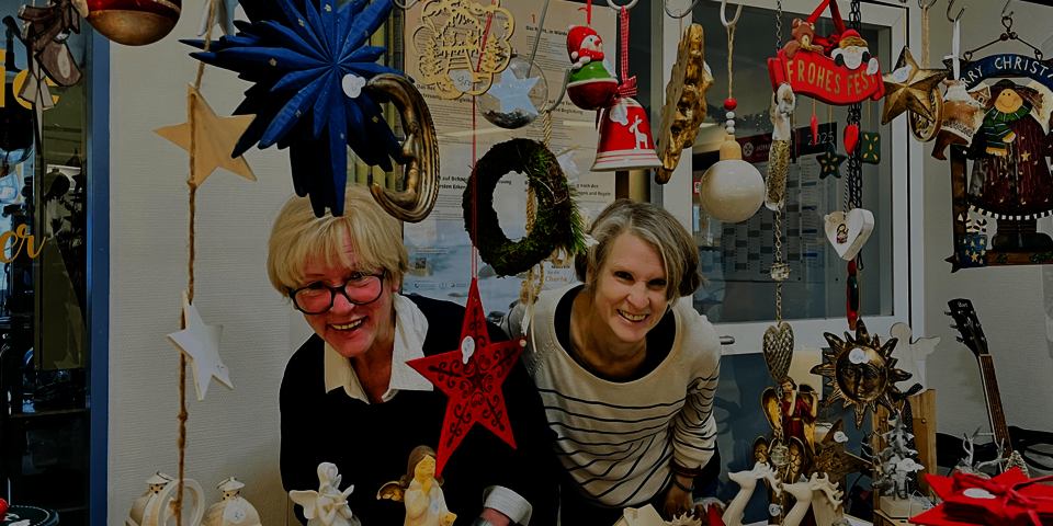 Zwei Frauen stehen hinter einem bunt geschmückten Weihnachtstisch mit vielen handgefertigten Dekoartikeln wie Sternen, Engeln, Kerzen und Weihnachtsfiguren, eingerahmt von hängenden Dekorationen.