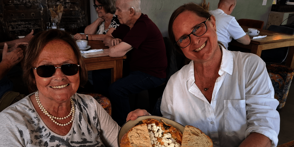 zwei Frauen, die jüngere präsentiert der Kamera eine dekorierten Teller mit Essen. Im Hintergrund weitere Personen an Tischen