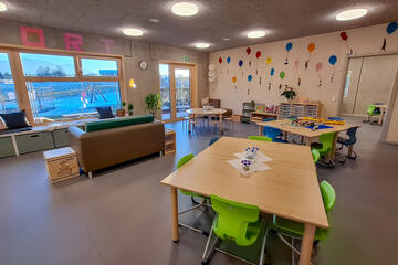 Heller Hort-Raum mit Tischen, bunten Stühlen, Sofa und großen Fenstern.