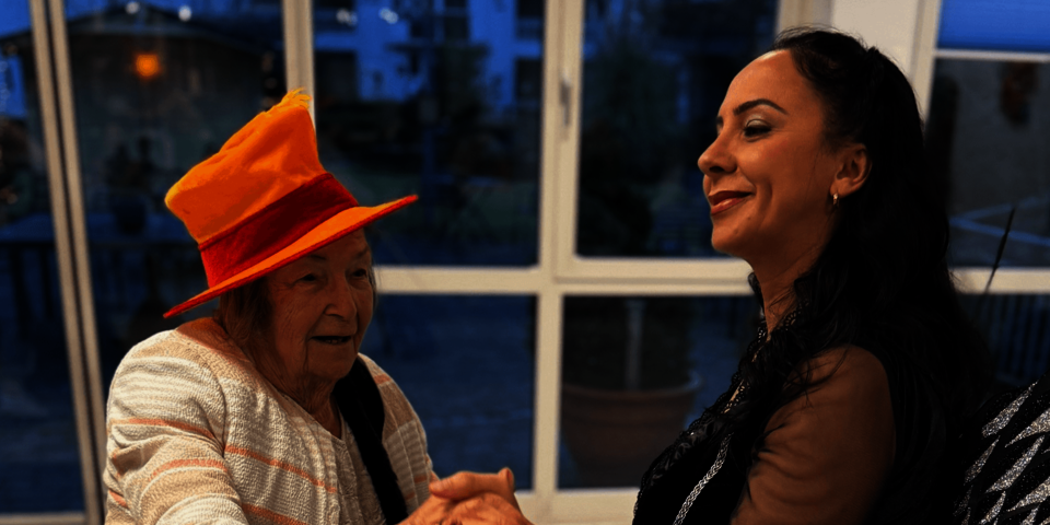 Ältere Person mit buntem Hut hält Händchen und tanzt mit einer lächelnden Frau in schwarzem Kostüm.