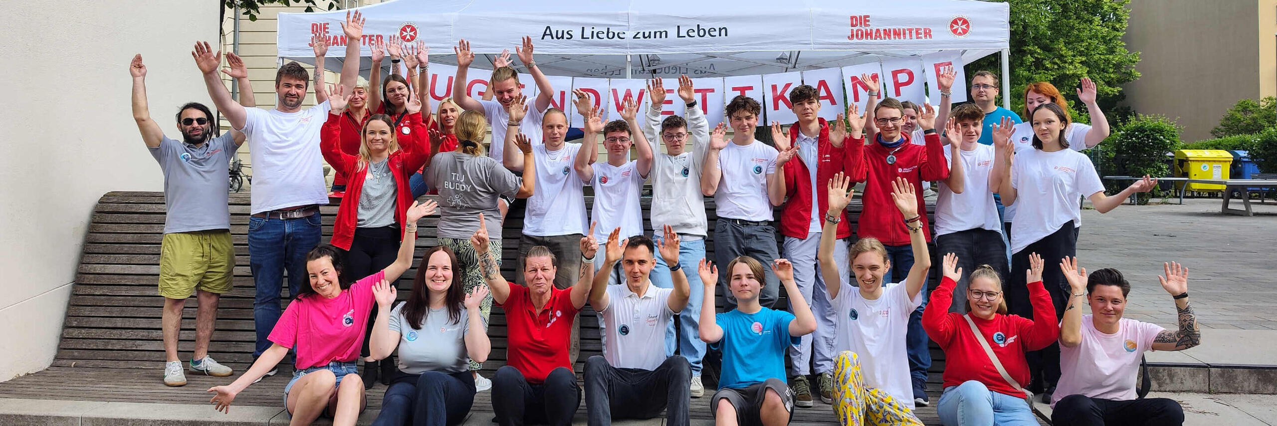Eine größere Gruppe von Jugendlichen und Mitarbeitenden der Johanniter-Jugend posiert auf einer Treppe vor einem Pavillon für ein Foto und streckt die Hände nach oben.