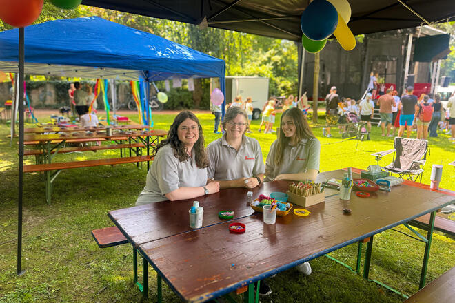 Drei Johanniterinnen sitzen an einem Tisch mit Bastelutensilien. Im Hintergrund ein Park mit Menschen