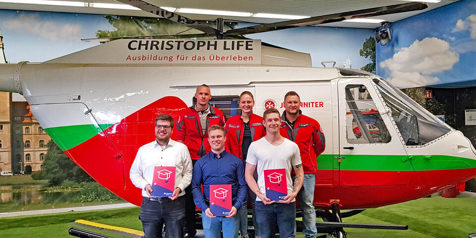 Drei junge Männer halten ein Diplom in der hand. Hinter ihnen stehen zwei Johanniter und eine Johanniterin. Im Hintergrund ist der Übungshelikopter Christoph life aus der San-Arena der Akademie zu sehen.