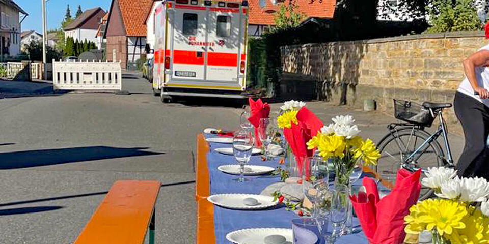 Eine lange schön eingedeckte Tafel auf einer Straße. Im Hintergrund ein RTW