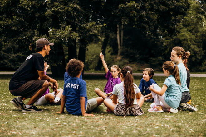 Kinder sitzen im Kreis auf der Wiese, ein Mädchen meldet sich, Betreuer hört zu.