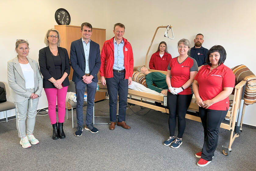 Philipp Tausch, Regionalvorstand der Johanniter in Südthüringen und Martin Bayer stehen mit Mitarbeiterinnen und Azubis im Johanniter-Pflegedienst Königsee an einem Pflegebett. Darin liegt die neue Puppe.