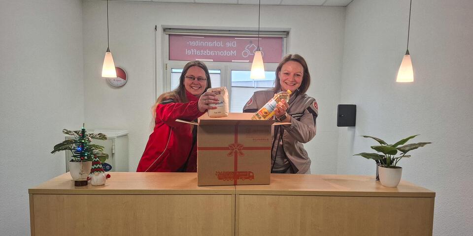 Zwei Frauen an einem Bürotresen. Vor ihnen steht ein offenes Paket und sie legen eine Flasche Öl und eine Packung Mehl hinein
