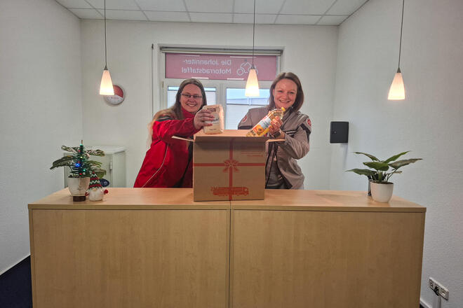 Zwei Frauen an einem Bürotresen. Vor ihnen steht ein offenes Paket und sie legen eine Flasche Öl und eine Packung Mehl hinein