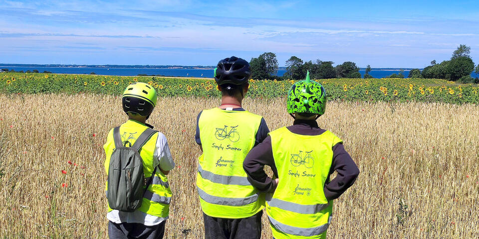 Drei Kinder stehen mit Fahrradhelm und Warnweste vor einer Wiese und schauen auf den Horizont. Man sieht das Meer.