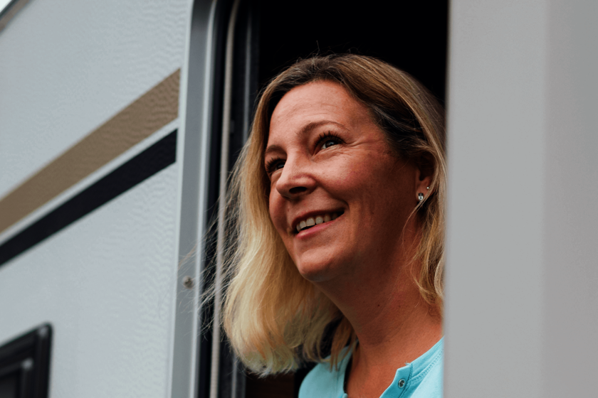 Nicole Eine blonde Frau in türkisem T-Shirt lächelt aus der Tür eines Wohnwagens.
