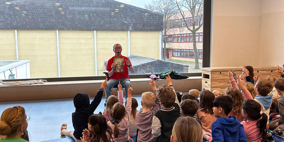 Ein Mann hält ein Buch und sitzt vor einer Gruppe von Kindern und Erwachsenen