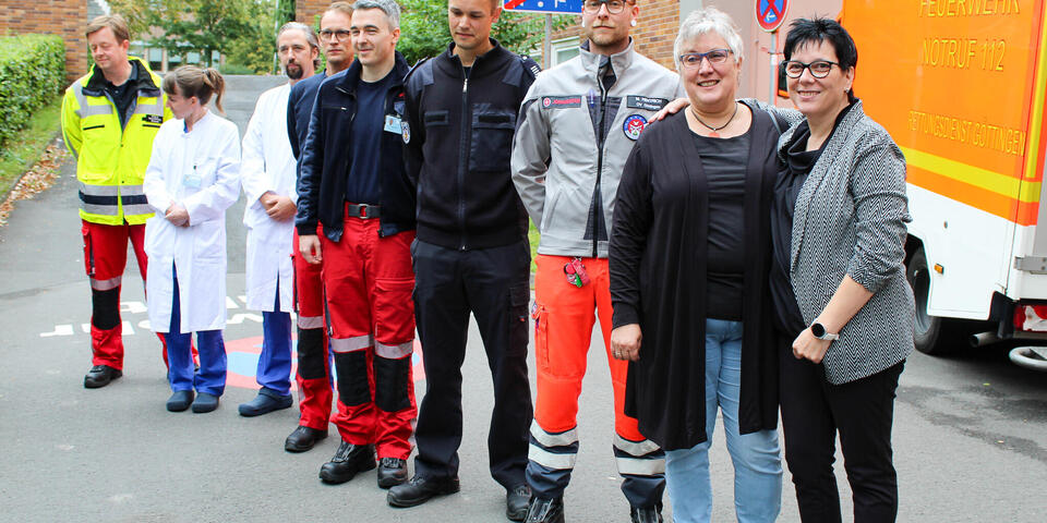 Zwei Frauen, mehrere Männer in Rettungsdienstbekleidung und ein Mann und eine Frau in Arztkitteln stehen vor einem Rettungswagen.